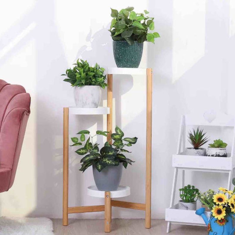 A three-tier wooden plant stand with white shelves, styled with potted green plants in a bright indoor setting next to a pink armchair and a white side table.