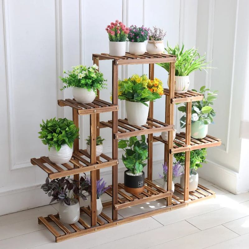 Multi-tiered brown wooden plant stand holding an assortment of potted green houseplants and colorful flowers, displayed indoors against a white paneled wall.