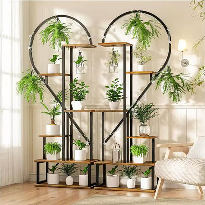 A large decorative metal plant stand shaped like two interlocking heart halves, with multiple wooden shelves and hanging green plants, shown in a bright living room next to a chair.