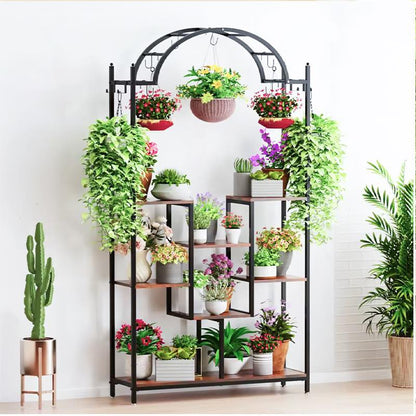 A black metal arched shelving unit with multiple tiers and hanging hooks, styled with numerous potted green plants and flowers, set against a white wall.